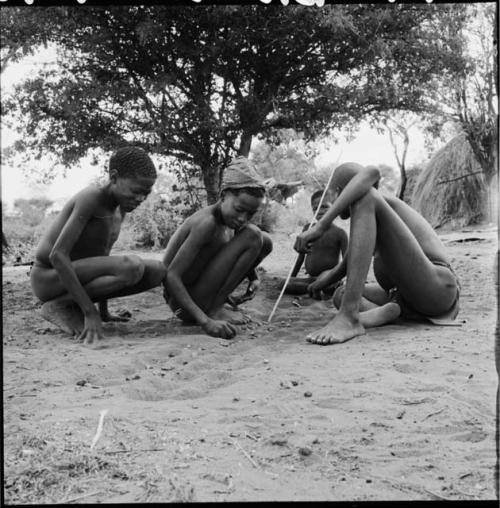 Four boys sitting, playing /Ui (the counting game), with the boy on the right pushing pebbles with a stick