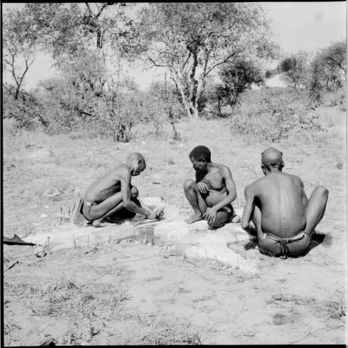 Man scraping a hide, sitting with ≠Toma and another man