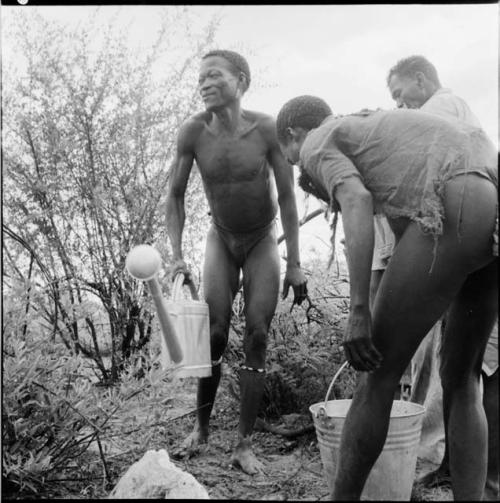 /Ti!kay carrying a watering can next to a man carrying a galvanized bucket, with Wilhelm Camm standing in the background