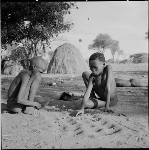 Two boys playing /Ui (the counting game) in the sand, with a skerm in the background