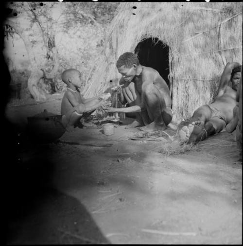 Boy squatting in front of a skerm with a man, showing him something and talking to him, with another man lying down next to the skerm