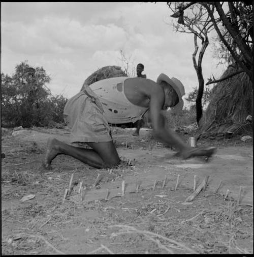 Man wearing a T-shirt, shorts and hat, leaning over, scraping an animal skin that is pegged to the ground