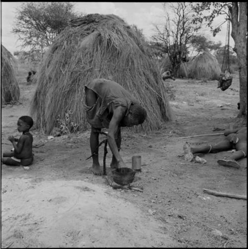 Man wearing a shirt, leaning over to reach his hand into a pot cooking on a fire, with a boy sitting near him, skerms in the background, meat hanging in a net bag from a nearby tree