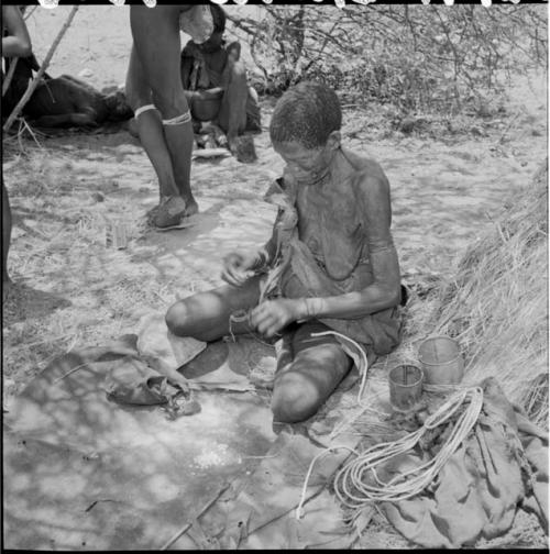 Woman sitting, stringing ostrich eggshell beads, with finished necklaces and a pile of beads on a kaross on the ground next to her