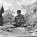 Woman sitting, stringing ostrich eggshell beads, with finished necklaces and a pile of beads on a kaross on the ground next to her