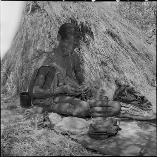 Woman sitting next to a skerm, stringing ostrich eggshell beads, with finished necklaces and a pile of beads on a kaross on the ground next to her