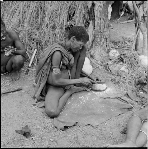 Woman wearing a safety pin in her ear, cutting a piece of ostrich eggshell with a knife, with ≠Toma sitting in the background eating