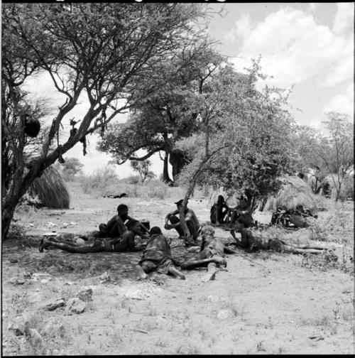 Group of men sitting and lying down in the shade, with skerms in the background