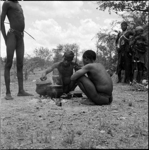 ≠Toma and another man cooking meat in two pots on a fire, with a man holding a bow standing behind them