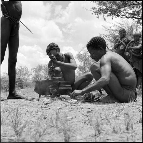 Man stirring a pot of meat cooking over a fire, with ≠Toma sitting next to him holding a plate of meat, a man holding a bow standing near them