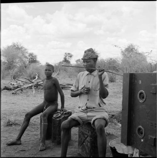 /Gaishay wearing Western clothing, sitting on an oil can, playing a one-stringed bow, with a boy sitting on a jerry can next to him