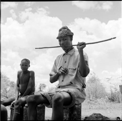 /Gaishay wearing Western clothing, sitting on an oil can, playing a one-stringed bow, with a boy sitting on a jerry can next to him