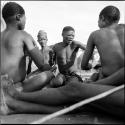 Four men sitting, including /Gunda, playing !ó'm!hú / !’Om g!xuni / ’Ai kota !U!u g!xuni / N!aih //’an (the porcupine game, also known as the axe game, assegai game or war game)