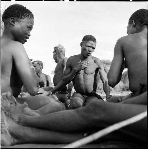 Four men sitting, including /Gunda, playing !ó'm!hú / !’Om g!xuni / ’Ai kota !U!u g!xuni / N!aih //’an (the porcupine game, also known as the axe game, assegai game or war game)