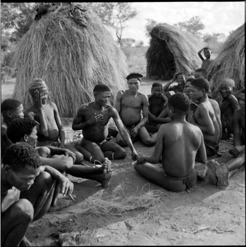 Group of men sitting, playing !ó'm!hú / !’Om g!xuni / ’Ai kota !U!u g!xuni / N!aih //’an (the porcupine game, also known as the axe game, assegai game or war game), with skerms in the background