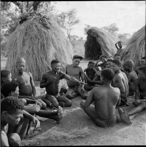 Group of men sitting, playing !ó'm!hú / !’Om g!xuni / ’Ai kota !U!u g!xuni / N!aih //’an (the porcupine game, also known as the axe game, assegai game or war game), with skerms in the background