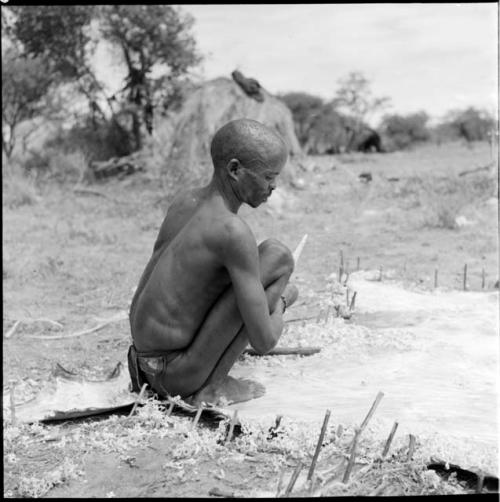 "/Gao Music" squatting on a hide pegged to the ground, holding a knife