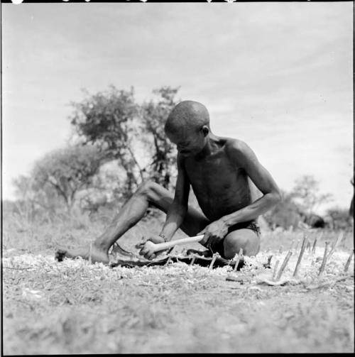 "/Gao Music" sits on a hide pegged to the ground, scraping it with his adze