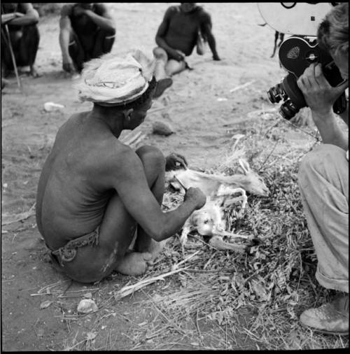 ≠Toma wearing a skin hat, sitting, skinning a small buck, with John Marshall filming him, view from behind