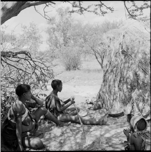 Woman preparing food with a digging stick, sitting with another woman outside a shelter skerm, with a woman lying on the ground, eating near them