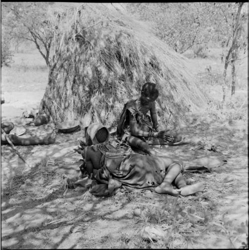 Woman sitting on a kaross next to a shelter skerm drilling ostrich eggshell beads, with another woman lying on the ground next to her