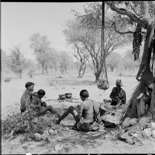 Group of men sitting next to a skerm with ostrich eggshells on the ground next to them, net bag and strips of meat hanging in a tree near them, two pots cooking on a fire behind them