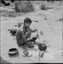 /Gunda stirring food in an iron pot with a stick, with a plate of food, broken clay pot, lid and tin cans on the ground next to him, group of people sitting in the background