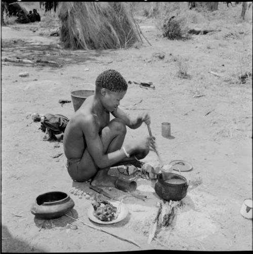 /Gunda stirring food in an iron pot with a stick, with a plate of food, broken clay pot, lid and tin cans on the ground next to him, group of people sitting in the background