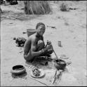 /Gunda stirring food in an iron pot with a stick, with a plate of food, broken clay pot, lid and tin cans on the ground next to him