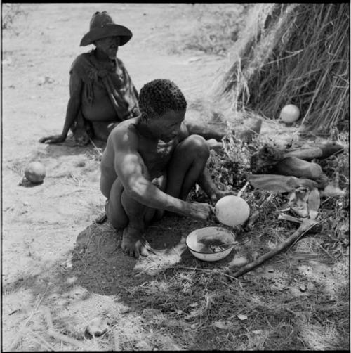 Man squatting, pulling out the plug in his //’ai [ostrich eggshell water container], with a man wearing a hat and a kaross tied around his shoulders sitting in the background