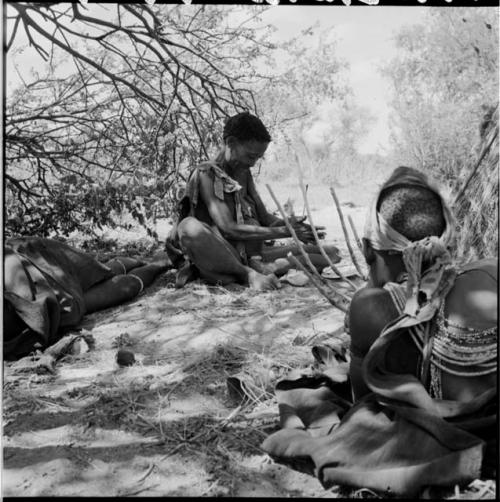 Woman sitting, holding a //guashi, view from behind, with a woman sitting, drilling holes in ostrich eggshell beads in the background