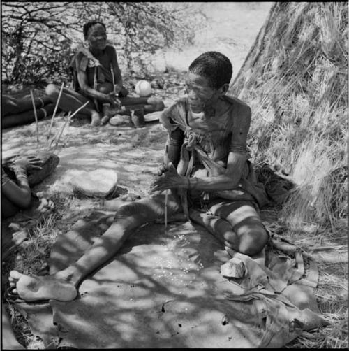 Two women sitting, drilling holes in ostrich eggshell beads, with a woman sitting and holding a //guashi near them