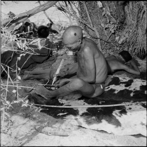 "Gao Medicine" sitting on a hide, holding a piece of sinew with his toes, working on it with a wooden knife, with a bow lying on the ground next to him