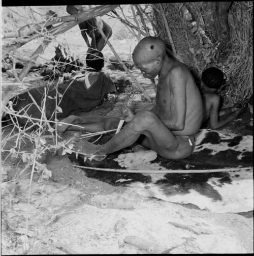 "Gao Medicine" sitting on a hide, holding a piece of sinew with his toes, working on it with a wooden knife, with a bow lying on the ground next to him