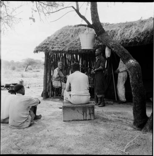 Men sitting with expedition members, including Laurence Marshall, in front of the expedition hut, with a galvanized bucket hanging in a tree near them