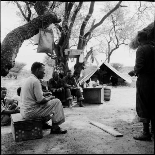 Men sitting with expedition members next to a table with food on it in the expedition camp, with belongings hanging in a tree near them