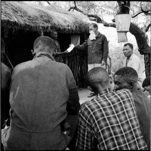 Group of expedition members sitting next to the expedition hut, one standing, wearing a fake beard