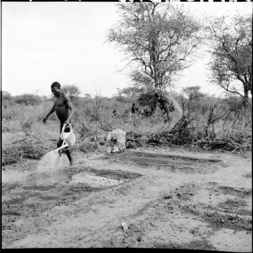 /Ti!kay watering plowed land with a watering can, with people sitting next to a skerm in the background