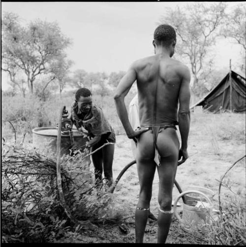 "/Gao Music" pumping water from a water drum into a watering can, with /Ti!kay standing, watching him, expedition tent in the background