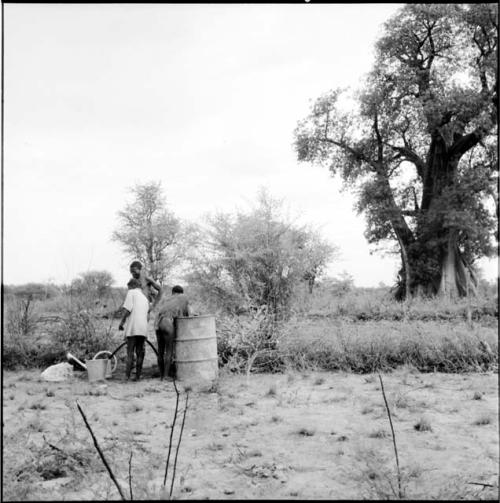 Three men standing next to a water drum, one filling a watering can with a hose