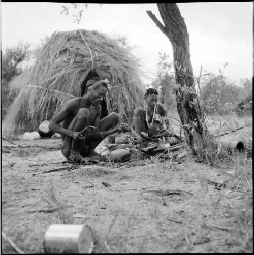 Man squatting, working on a piece of sinew, with a woman sitting next to him, skerm in the background