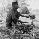 Man squatting, holding a piece of meat and a knife, with an iron pot full of food sitting in the ashes of a fire, meat placed on leaves next to him