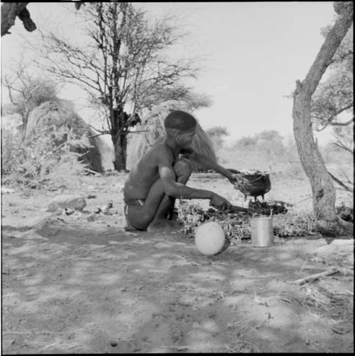 Man squatting, holding a piece of meat and a knife, with an iron pot full of food sitting in the ashes of a fire, meat placed on leaves and an ostrich eggshell next to him