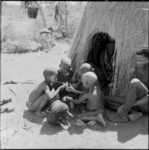 "/Qui Umbilicus" using a toothpick, sitting outside his skerm next to four boys eating out of a dish next to an iron pot, with a woman sitting inside the skerm behind them