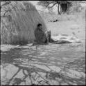 Man sitting, leaning against a skerm, a hide pegged to the ground near him, a dried hide on the other side of him