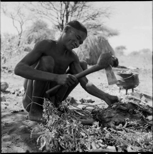 Man chopping meat with an axe, with a cooking pot on the ground behind him, skerm in the background