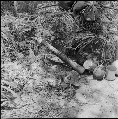 Possessions on and next to a bush, including hunting bags, bow, quiver, two tin cans and a melon