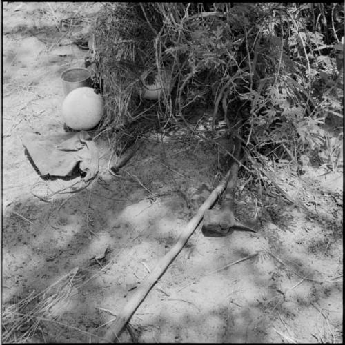 Possessions on the ground next to a bush, including an axe, stick, eggshell and tin cup