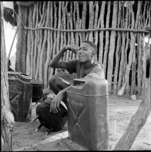 Man wearing a T-shirt, sitting next to the expedition hut, eating from an iron pot, with two jerry cans on the ground next to him