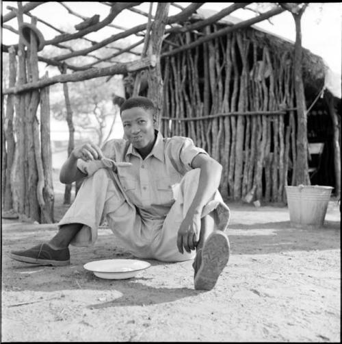 Man wearing Western clothes sitting outside the expedition hut, eating from an enamel dish